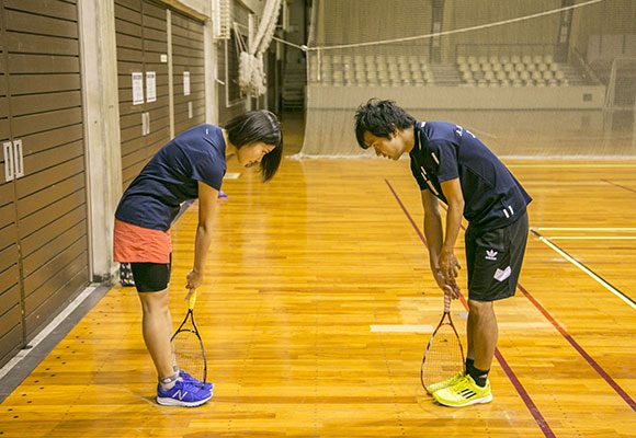 足の間にラケットを挟み、床と垂直に立てる。利き腕を胸の前に伸ばし、握手をするように手のひらを内側に向け、そのままラケットを握る。